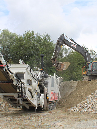 Bagger lädt Abbruchmaterial in den Trichter des Brechers. Vorne ist das gebrochene und gesiebte Produkt zu sehen.